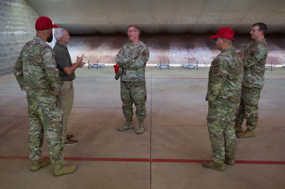 Five individuals are gathered in what appears to be a large indoor shooting range or hardened aircraft shelter, engaged in conversation. Four are wearing military camouflage uniforms (OCP pattern), with two wearing red baseball caps, while one civilian in a green polo shirt and khaki pants participates in the discussion. The central figure stands with arms crossed while others gesture during what appears to be a briefing or informal discussion.