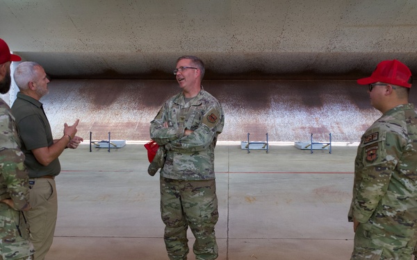 Five individuals are gathered in what appears to be a large indoor shooting range or hardened aircraft shelter, engaged in conversation. Four are wearing military camouflage uniforms (OCP pattern), with two wearing red baseball caps, while one civilian in a green polo shirt and khaki pants participates in the discussion. The central figure stands with arms crossed while others gesture during what appears to be a briefing or informal discussion.