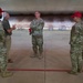 Five individuals are gathered in what appears to be a large indoor shooting range or hardened aircraft shelter, engaged in conversation. Four are wearing military camouflage uniforms (OCP pattern), with two wearing red baseball caps, while one civilian in a green polo shirt and khaki pants participates in the discussion. The central figure stands with arms crossed while others gesture during what appears to be a briefing or informal discussion.