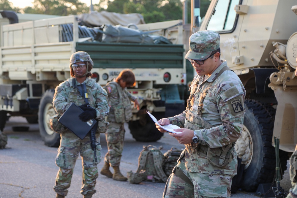 The image shows U.S. Army soldiers in Operational Camouflage Pattern (OCP) uniforms gathered near military tactical vehicles, likely during a staging or pre-mission briefing. One soldier in the foreground is reviewing documents while another stands holding a clipboard or folder. The setting appears to be a motor pool or staging area during early morning or evening hours.