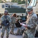 The image shows U.S. Army soldiers in Operational Camouflage Pattern (OCP) uniforms gathered near military tactical vehicles, likely during a staging or pre-mission briefing. One soldier in the foreground is reviewing documents while another stands holding a clipboard or folder. The setting appears to be a motor pool or staging area during early morning or evening hours.