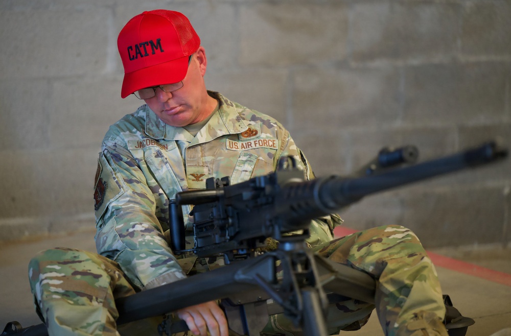 A U.S. Air Force personnel member wearing OCP (Operational Camouflage Pattern) uniform is seated and examining a large crew-served weapon, likely an M240 machine gun. The individual is identified by name tape as 'Jacobson' and wears a red 'CATM' (Combat Arms Training and Maintenance) baseball cap, indicating a weapons instructor or trainer role. The setting appears to be an indoor range or armory facility with concrete block walls.