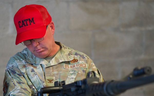 A U.S. Air Force personnel member wearing OCP (Operational Camouflage Pattern) uniform is seated and examining a large crew-served weapon, likely an M240 machine gun. The individual is identified by name tape as 'Jacobson' and wears a red 'CATM' (Combat Arms Training and Maintenance) baseball cap, indicating a weapons instructor or trainer role. The setting appears to be an indoor range or armory facility with concrete block walls.