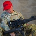 A U.S. Air Force personnel member wearing OCP (Operational Camouflage Pattern) uniform is seated and examining a large crew-served weapon, likely an M240 machine gun. The individual is identified by name tape as 'Jacobson' and wears a red 'CATM' (Combat Arms Training and Maintenance) baseball cap, indicating a weapons instructor or trainer role. The setting appears to be an indoor range or armory facility with concrete block walls.