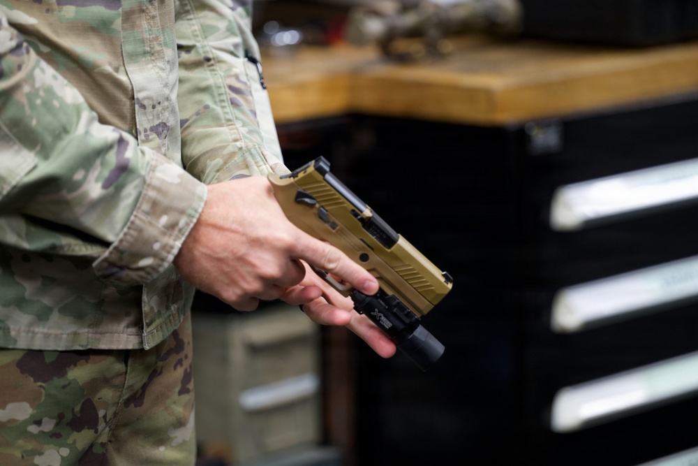 A person wearing military OCP (Operational Camouflage Pattern) uniform is holding a tan/flat dark earth semi-automatic pistol, consistent with a SIG Sauer P320 M17/M18, with a weapon-mounted light attached to the rail. The setting appears to be an armory or maintenance workspace, indicated by the workbench and tool chest visible in the background. The firearm is being held in a two-handed administrative grip, not in a firing stance.