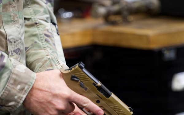 A person wearing military OCP (Operational Camouflage Pattern) uniform is holding a tan/flat dark earth semi-automatic pistol, consistent with a SIG Sauer P320 M17/M18, with a weapon-mounted light attached to the rail. The setting appears to be an armory or maintenance workspace, indicated by the workbench and tool chest visible in the background. The firearm is being held in a two-handed administrative grip, not in a firing stance.