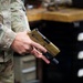 A person wearing military OCP (Operational Camouflage Pattern) uniform is holding a tan/flat dark earth semi-automatic pistol, consistent with a SIG Sauer P320 M17/M18, with a weapon-mounted light attached to the rail. The setting appears to be an armory or maintenance workspace, indicated by the workbench and tool chest visible in the background. The firearm is being held in a two-handed administrative grip, not in a firing stance.