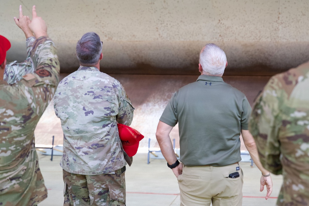 The image shows a group of individuals, some in military camouflage uniforms and one in civilian attire, viewed from behind. They appear to be receiving a briefing or tour near a large structure, possibly an aircraft or military installation. One person in uniform is gesturing upward, apparently pointing out or explaining something.