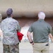 The image shows a group of individuals, some in military camouflage uniforms and one in civilian attire, viewed from behind. They appear to be receiving a briefing or tour near a large structure, possibly an aircraft or military installation. One person in uniform is gesturing upward, apparently pointing out or explaining something.