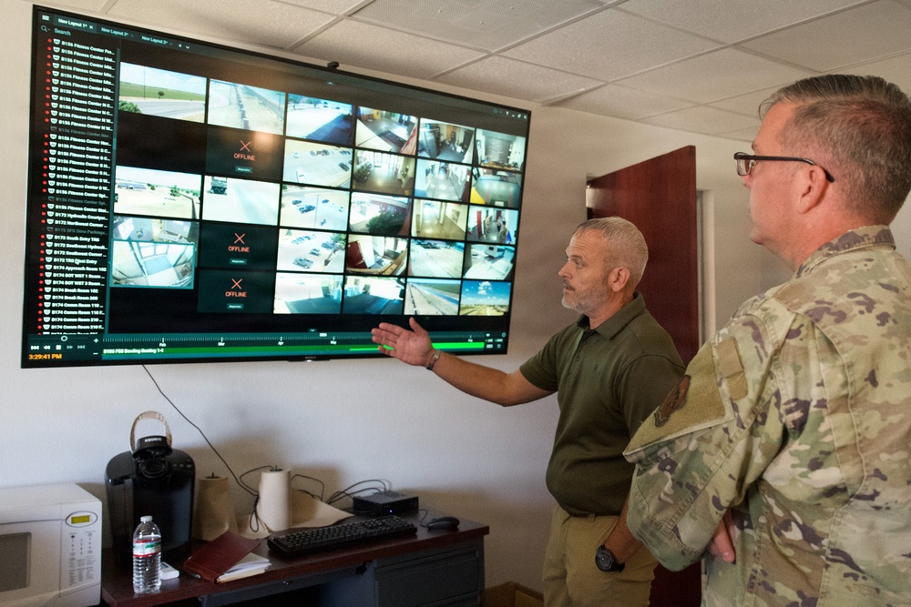 Two individuals are reviewing a large wall-mounted security surveillance monitor displaying multiple camera feeds. One person in a green polo shirt is gesturing toward the screen while explaining something to a person in military camouflage uniform. Several camera feeds show 'OFFLINE' status, while others display various indoor and outdoor locations.