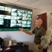 Two individuals are reviewing a large wall-mounted security surveillance monitor displaying multiple camera feeds. One person in a green polo shirt is gesturing toward the screen while explaining something to a person in military camouflage uniform. Several camera feeds show 'OFFLINE' status, while others display various indoor and outdoor locations.