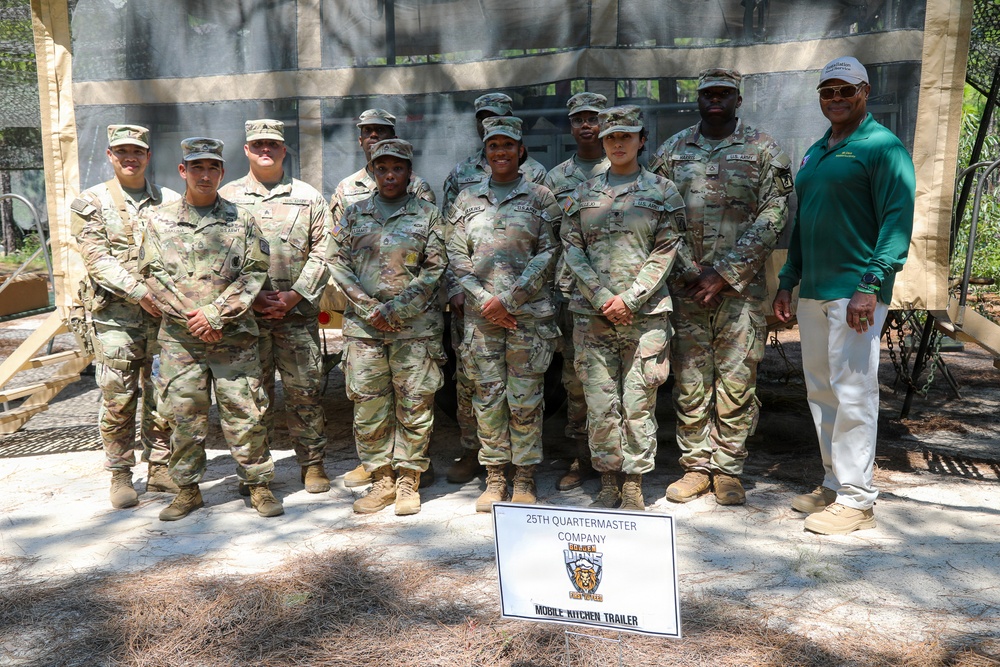 A group photo of approximately ten U.S. Army soldiers in OCP (Operational Camouflage Pattern) uniforms posing with one civilian in a green polo shirt and white pants. They are standing in front of a field shelter structure with a sign identifying them as the 25th Quartermaster Company Mobile Kitchen Trailer unit. The setting appears to be an outdoor military training or demonstration area.
