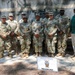A group photo of approximately ten U.S. Army soldiers in OCP (Operational Camouflage Pattern) uniforms posing with one civilian in a green polo shirt and white pants. They are standing in front of a field shelter structure with a sign identifying them as the 25th Quartermaster Company Mobile Kitchen Trailer unit. The setting appears to be an outdoor military training or demonstration area.