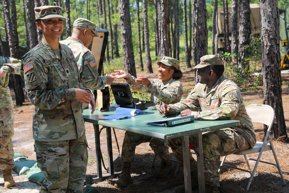 A group of U.S. Army soldiers in OCP (Operational Camouflage Pattern) uniforms are gathered at an outdoor field station set up among pine trees. A senior officer (identifiable by a lieutenant colonel oak leaf cluster on her patrol cap) is interacting with soldiers seated at a folding table with documents and a tablet. The setting appears to be a field training exercise or operational briefing area.