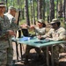 A group of U.S. Army soldiers in OCP (Operational Camouflage Pattern) uniforms are gathered at an outdoor field station set up among pine trees. A senior officer (identifiable by a lieutenant colonel oak leaf cluster on her patrol cap) is interacting with soldiers seated at a folding table with documents and a tablet. The setting appears to be a field training exercise or operational briefing area.