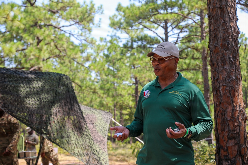 A man identified as 'Mr. Clark' from 'Installation Food Service' is speaking or presenting outdoors in a wooded military training area. He is wearing a green long-sleeve polo shirt with patches and a name tag, a cap reading 'Installation Food Service,' and yellow-tinted sunglasses. In the background, soldiers in camouflage uniforms are visible near camouflage netting strung between trees.