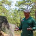 A man identified as 'Mr. Clark' from 'Installation Food Service' is speaking or presenting outdoors in a wooded military training area. He is wearing a green long-sleeve polo shirt with patches and a name tag, a cap reading 'Installation Food Service,' and yellow-tinted sunglasses. In the background, soldiers in camouflage uniforms are visible near camouflage netting strung between trees.