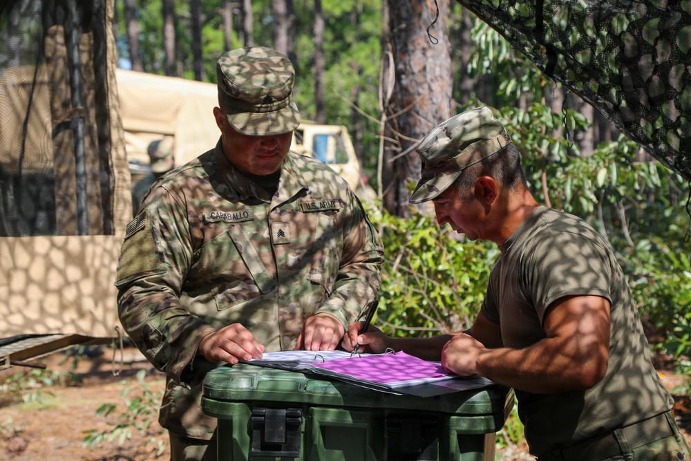 Two U.S. Army soldiers are reviewing documents at a field operations site in a wooded area. The soldier on the left, identified by his name tape as 'Caraballo' with Sergeant rank insignia, is in full OCP uniform, while the soldier on the right wears a camouflage cap and olive t-shirt. They appear to be conducting administrative or operational planning tasks at a forward operating base or field training exercise.