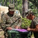 Two U.S. Army soldiers are reviewing documents at a field operations site in a wooded area. The soldier on the left, identified by his name tape as 'Caraballo' with Sergeant rank insignia, is in full OCP uniform, while the soldier on the right wears a camouflage cap and olive t-shirt. They appear to be conducting administrative or operational planning tasks at a forward operating base or field training exercise.