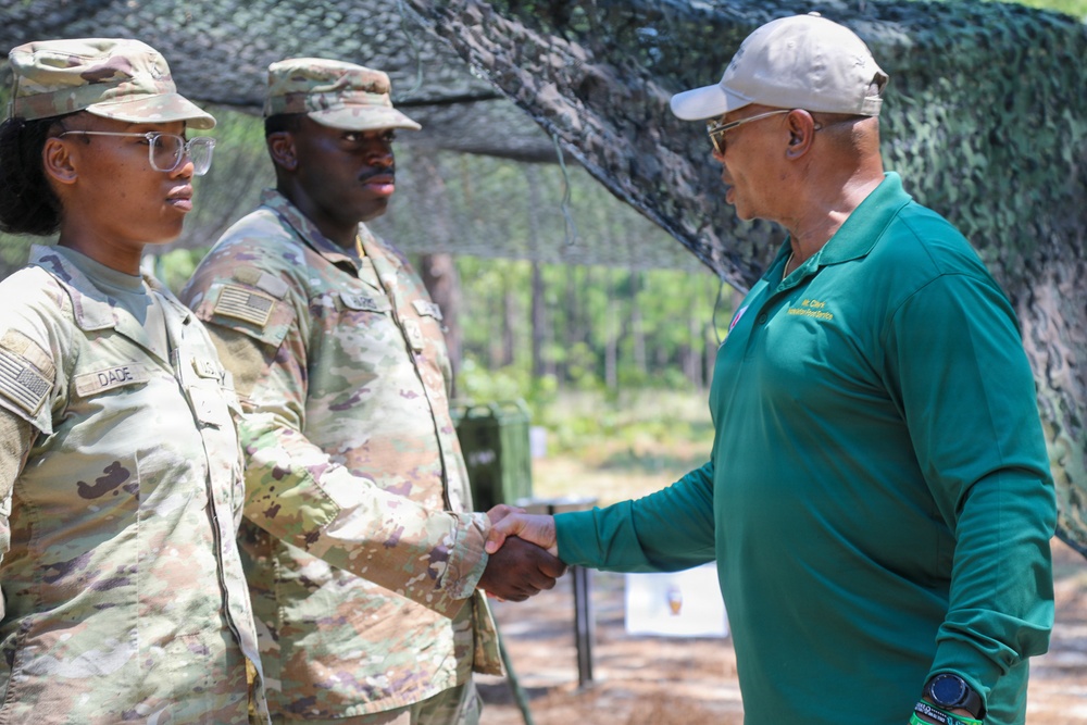A civilian man in a green polo shirt and white cap is shaking hands with a female U.S. Army soldier in OCP (Operational Camouflage Pattern) uniform, whose name tape reads 'DADE'. A second soldier in OCP uniform stands in the background. The setting appears to be an outdoor military field environment with camouflage netting overhead.