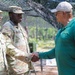 A civilian man in a green polo shirt and white cap is shaking hands with a female U.S. Army soldier in OCP (Operational Camouflage Pattern) uniform, whose name tape reads 'DADE'. A second soldier in OCP uniform stands in the background. The setting appears to be an outdoor military field environment with camouflage netting overhead.
