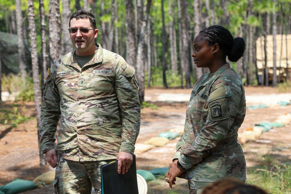 Two U.S. Army soldiers in OCP (Operational Camouflage Pattern) uniforms are standing outdoors in a wooded training area. The male soldier on the left, whose name tape reads 'MILLS,' appears to be speaking, while the female soldier on the right listens attentively. The setting suggests a field training exercise or outdoor briefing environment.