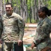 Two U.S. Army soldiers in OCP (Operational Camouflage Pattern) uniforms are standing outdoors in a wooded training area. The male soldier on the left, whose name tape reads 'MILLS,' appears to be speaking, while the female soldier on the right listens attentively. The setting suggests a field training exercise or outdoor briefing environment.
