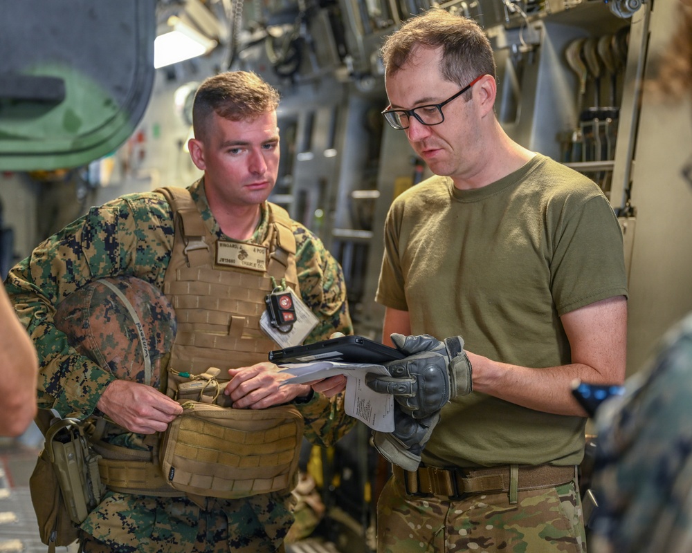 Two military personnel are reviewing documents or a tablet inside what appears to be a military aircraft cargo hold. The individual on the left is wearing USMC MARPAT camouflage with a plate carrier vest, while the individual on the right wears an olive drab t-shirt with MultiCam trousers and tactical gloves. They appear to be conducting a briefing, inspection, or load planning review.
