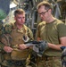 Two military personnel are reviewing documents or a tablet inside what appears to be a military aircraft cargo hold. The individual on the left is wearing USMC MARPAT camouflage with a plate carrier vest, while the individual on the right wears an olive drab t-shirt with MultiCam trousers and tactical gloves. They appear to be conducting a briefing, inspection, or load planning review.