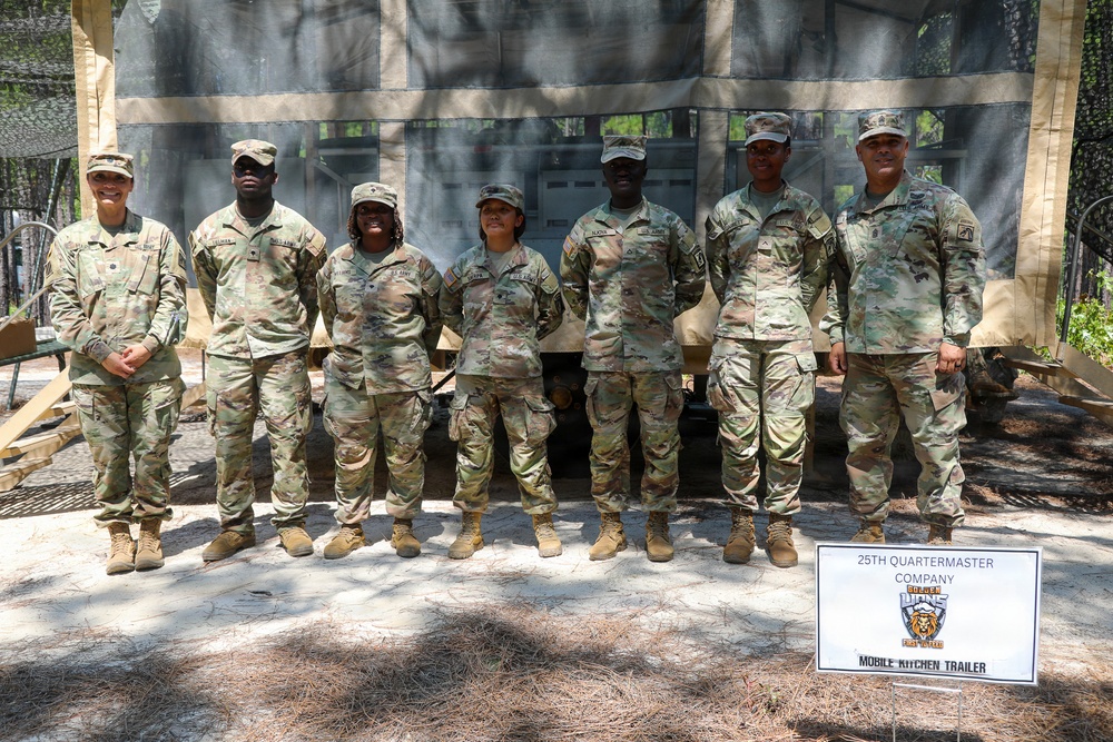 Seven U.S. Army soldiers in OCP (Operational Camouflage Pattern) uniforms pose together in front of a Mobile Kitchen Trailer belonging to the 25th Quartermaster Company. The group is standing outdoors in a wooded area under a camouflage netting structure. A sign identifying the unit and equipment is visible in the lower right of the frame.