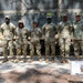 Seven U.S. Army soldiers in OCP (Operational Camouflage Pattern) uniforms pose together in front of a Mobile Kitchen Trailer belonging to the 25th Quartermaster Company. The group is standing outdoors in a wooded area under a camouflage netting structure. A sign identifying the unit and equipment is visible in the lower right of the frame.