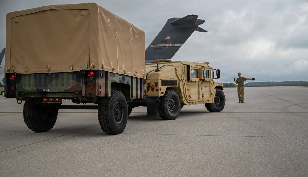 A military HMMWV (Humvee) towing a covered cargo trailer is positioned on an airfield tarmac near a large military transport aircraft. A service member in camouflage uniform is visible to the right, appearing to direct or signal the vehicle. The aircraft tail section bears 'AFRC' markings and the number '30804', indicating Air Force Reserve Command.