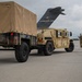 A military HMMWV (Humvee) towing a covered cargo trailer is positioned on an airfield tarmac near a large military transport aircraft. A service member in camouflage uniform is visible to the right, appearing to direct or signal the vehicle. The aircraft tail section bears 'AFRC' markings and the number '30804', indicating Air Force Reserve Command.
