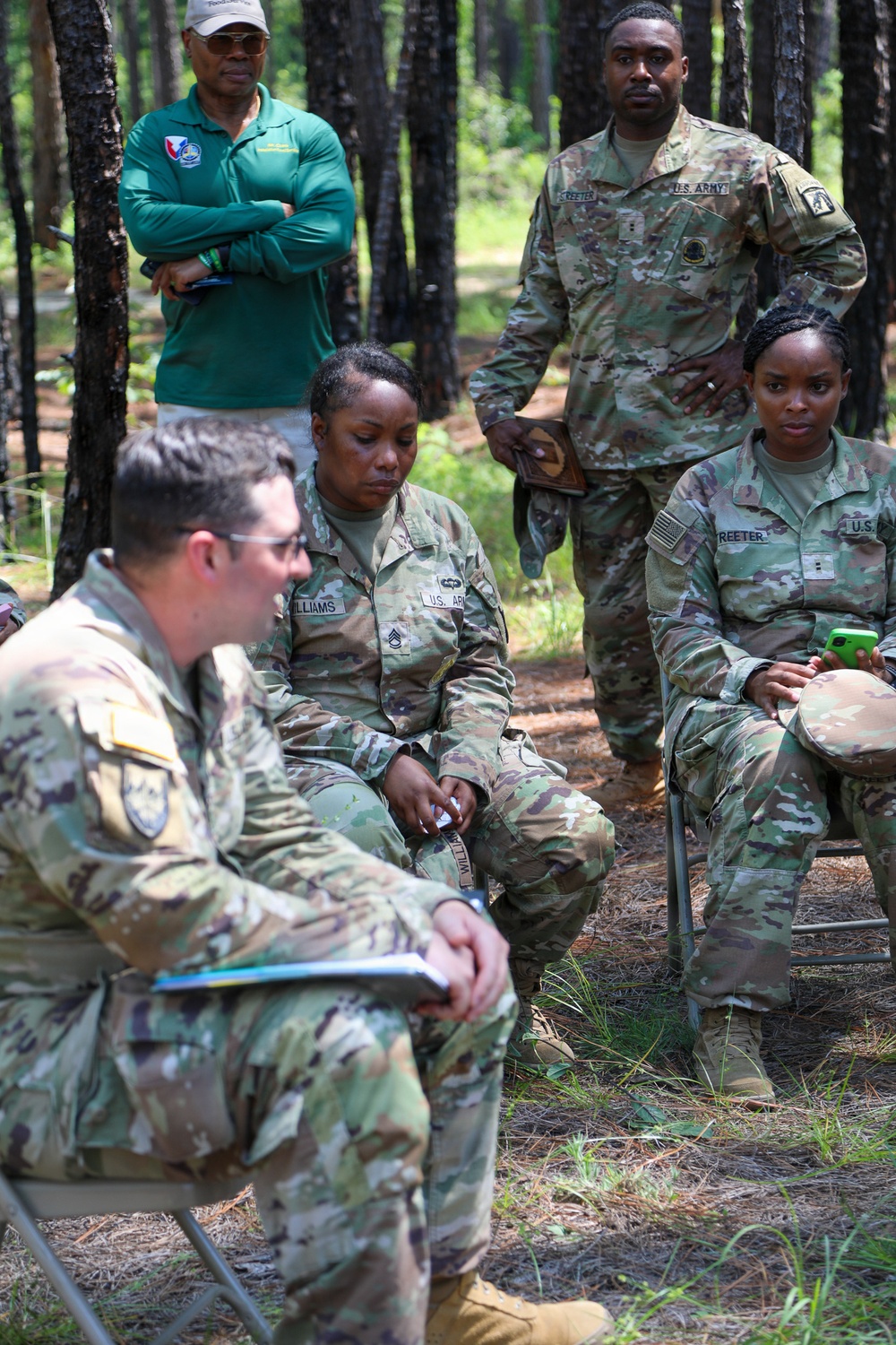 A group of U.S. Army soldiers in OCP (Operational Camouflage Pattern) uniforms are gathered in a wooded outdoor setting, appearing to be in a briefing or after-action review. A civilian in a green polo shirt stands in the background observing. Visible name tapes include 'Williams' and 'Reeter', and soldiers are seated and standing in an informal outdoor meeting configuration.