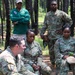 A group of U.S. Army soldiers in OCP (Operational Camouflage Pattern) uniforms are gathered in a wooded outdoor setting, appearing to be in a briefing or after-action review. A civilian in a green polo shirt stands in the background observing. Visible name tapes include 'Williams' and 'Reeter', and soldiers are seated and standing in an informal outdoor meeting configuration.