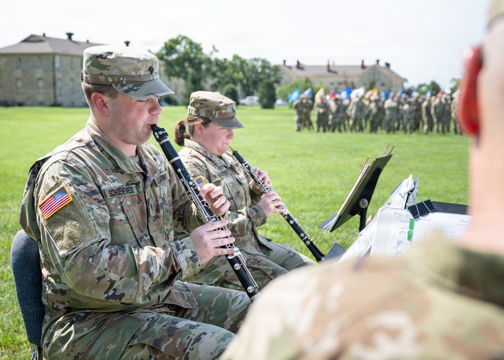 U.S. Army musicians in OCP (Operational Camouflage Pattern) uniforms are performing on clarinets during an outdoor military ceremony. The foreground shows at least two clearly visible soldiers playing clarinets while reading sheet music from stands. In the background, a formation of soldiers with color guard flags is assembled on a parade field flanked by historic stone buildings.