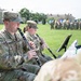 U.S. Army musicians in OCP (Operational Camouflage Pattern) uniforms are performing on clarinets during an outdoor military ceremony. The foreground shows at least two clearly visible soldiers playing clarinets while reading sheet music from stands. In the background, a formation of soldiers with color guard flags is assembled on a parade field flanked by historic stone buildings.
