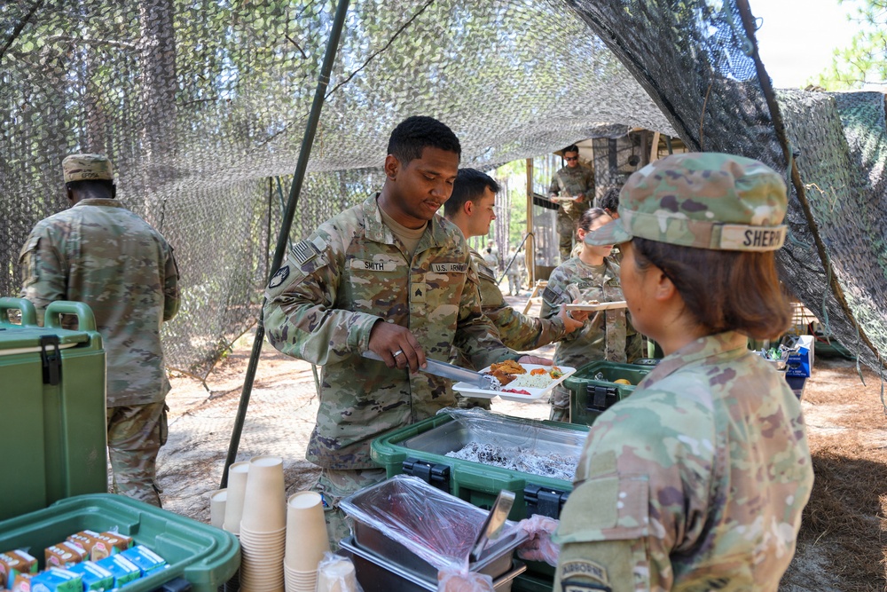 U.S. Army soldiers are participating in a field mess operation under a camouflage netting canopy in an outdoor wooded setting. Soldier 'Smith' is being served food by Soldier 'Sherm' at a field chow line with green insulated food containers. Multiple soldiers are visible in the background also receiving or preparing food.