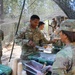 U.S. Army soldiers are participating in a field mess operation under a camouflage netting canopy in an outdoor wooded setting. Soldier 'Smith' is being served food by Soldier 'Sherm' at a field chow line with green insulated food containers. Multiple soldiers are visible in the background also receiving or preparing food.