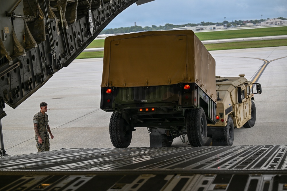 A military loading operation is underway at an airfield, where a HMMWV (Humvee) towing a covered trailer is being driven up the rear cargo ramp of a large military transport aircraft, likely a C-17 Globemaster III. A single service member in camouflage uniform stands to the left of the ramp, observing and guiding the loading process. The setting appears to be a domestic U.S. military airfield based on the visible runway infrastructure and surrounding vegetation.
