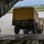 Air Force, Marines conduct joint training on C-17 A military loading operation is underway at an airfield, where a HMMWV (Humvee) towing a covered trailer is being driven up the rear cargo ramp of a large military transport aircraft, likely a C-17 Globemaster III. A single service member in camouflage uniform stands to the left of the ramp, observing and guiding the loading process. The setting appears to be a domestic U.S. military airfield based on the visible runway infrastructure and surrounding vegetation.