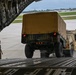 A military loading operation is underway at an airfield, where a HMMWV (Humvee) towing a covered trailer is being driven up the rear cargo ramp of a large military transport aircraft, likely a C-17 Globemaster III. A single service member in camouflage uniform stands to the left of the ramp, observing and guiding the loading process. The setting appears to be a domestic U.S. military airfield based on the visible runway infrastructure and surrounding vegetation.