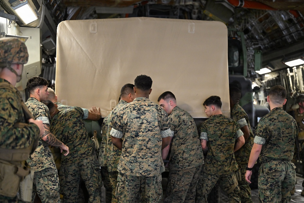 A group of military personnel in digital camouflage uniforms are working together inside what appears to be a military cargo aircraft. They are collectively pushing or maneuvering a large tan/beige covered object, likely a piece of equipment or cargo. The scene depicts a coordinated loading or unloading operation within the aircraft's cargo bay.