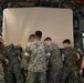 A group of military personnel in digital camouflage uniforms are working together inside what appears to be a military cargo aircraft. They are collectively pushing or maneuvering a large tan/beige covered object, likely a piece of equipment or cargo. The scene depicts a coordinated loading or unloading operation within the aircraft's cargo bay.
