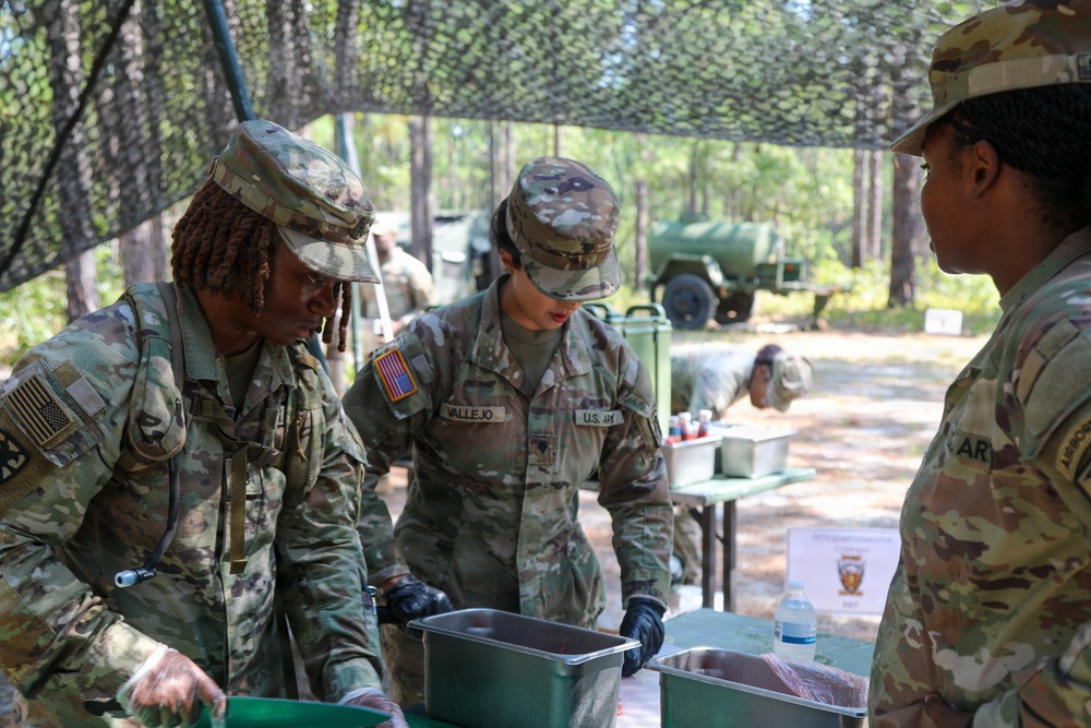 Three U.S. Army soldiers in OCP (Operational Camouflage Pattern) uniforms are working at an outdoor field station under camouflage netting. Two soldiers are actively handling food service containers while wearing black gloves, suggesting a field kitchen or food service operation. The setting appears to be a wooded training area with military vehicles visible in the background.
