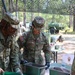 Three U.S. Army soldiers in OCP (Operational Camouflage Pattern) uniforms are working at an outdoor field station under camouflage netting. Two soldiers are actively handling food service containers while wearing black gloves, suggesting a field kitchen or food service operation. The setting appears to be a wooded training area with military vehicles visible in the background.