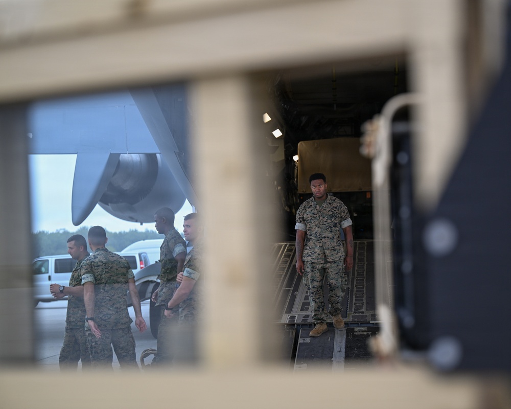 The image is photographed through a vehicle window or hatch frame, showing military personnel in camouflage uniforms on an airfield tarmac near a large military transport aircraft. One service member stands on the loading ramp of the aircraft while others congregate nearby. A military vehicle or equipment is visible inside the aircraft's cargo hold.