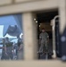 The image is photographed through a vehicle window or hatch frame, showing military personnel in camouflage uniforms on an airfield tarmac near a large military transport aircraft. One service member stands on the loading ramp of the aircraft while others congregate nearby. A military vehicle or equipment is visible inside the aircraft's cargo hold.
