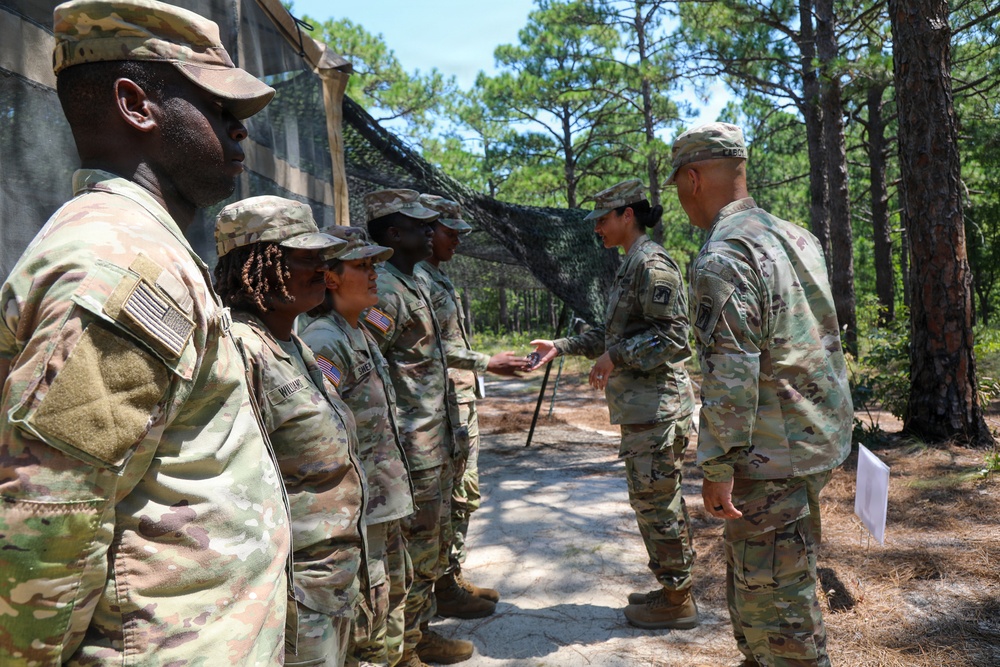 A group of U.S. Army soldiers in OCP (Operational Camouflage Pattern) uniforms are lined up in a wooded outdoor training area. A soldier appears to be handing or receiving an item from a senior soldier, suggesting an inspection, award ceremony, or briefing activity. The setting includes pine trees, camouflage netting, and a sandy path consistent with a military training installation.