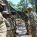 A group of U.S. Army soldiers in OCP (Operational Camouflage Pattern) uniforms are lined up in a wooded outdoor training area. A soldier appears to be handing or receiving an item from a senior soldier, suggesting an inspection, award ceremony, or briefing activity. The setting includes pine trees, camouflage netting, and a sandy path consistent with a military training installation.